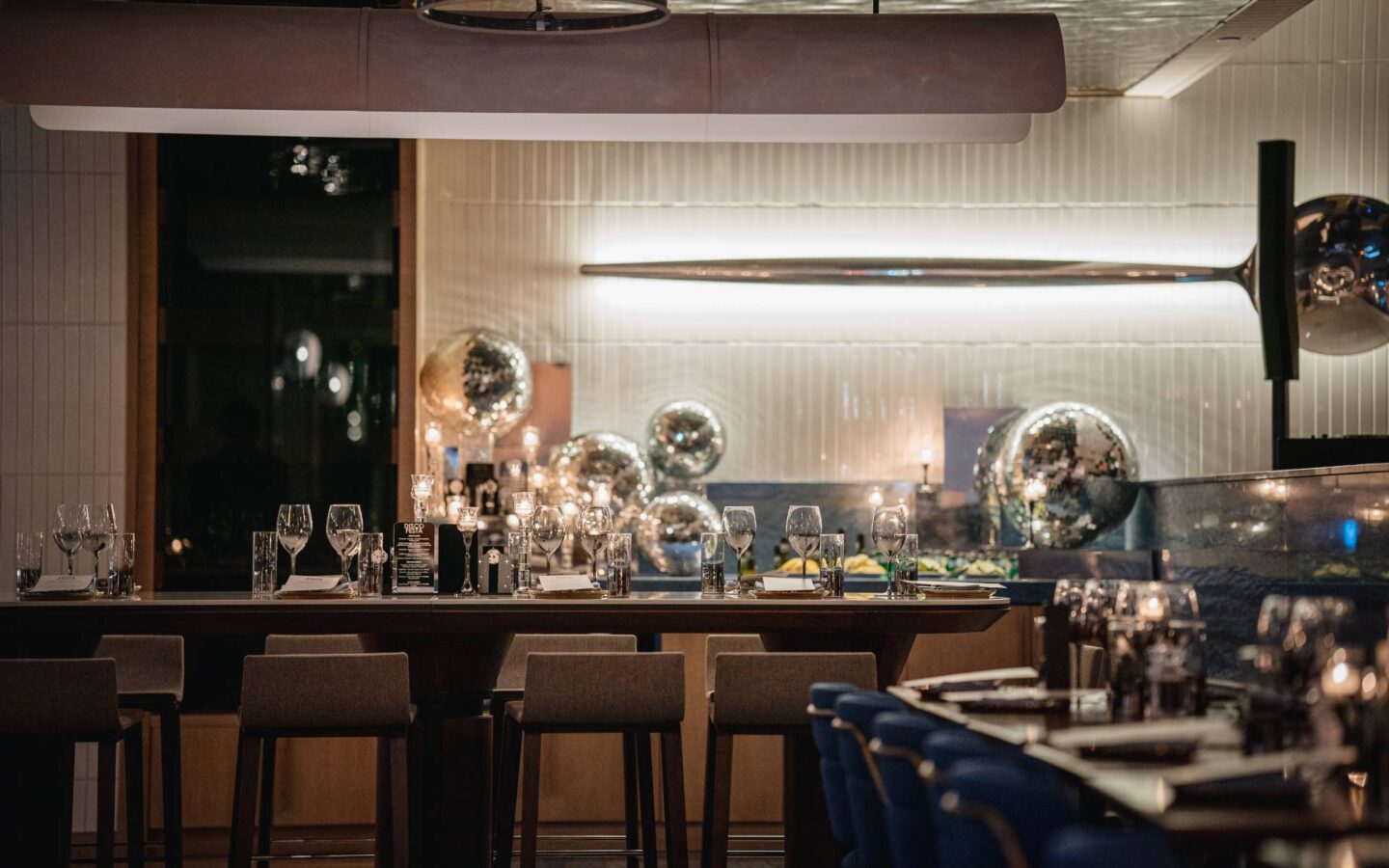 Modern restaurant dining room with high-top seating, elegantly set tables, ambient lighting, and reflective metallic décor creating a sophisticated evening atmosphere