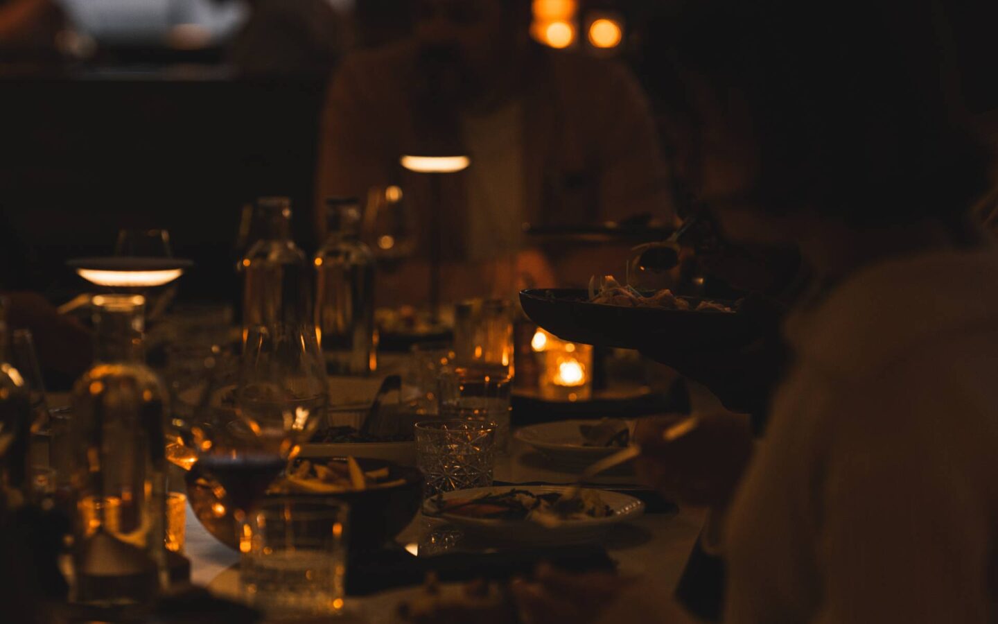 Warmly lit restaurant table with diners enjoying a candlelit meal as a server presents a dish, creating an intimate evening dining atmosphere.